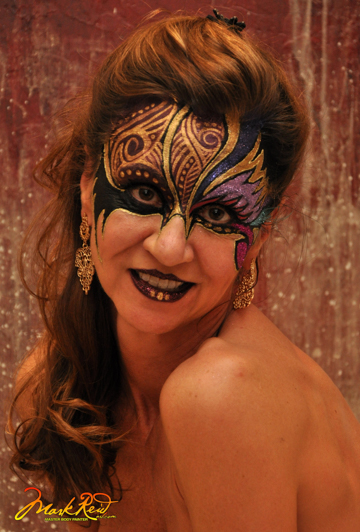 brunette woman with an elaborate uppfer face painting in purple, gold, and black